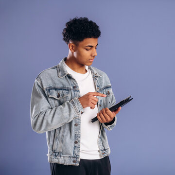 Young African American male wearing a denim jacket and white t-shirt holds a tablet while standing against a solid purple background