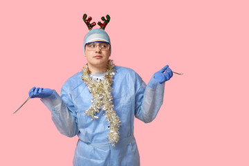 Portrait of young male surgeon with tools, Christmas tinsel and reindeer headband on pink background © Pixel-Shot
