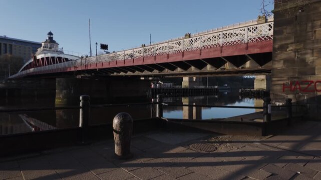 Ground-level pan of the historic Swing Bridge in Newcastle at dawn, highlighting the industrial iron structure and stone pillars under the low early morning sun.
