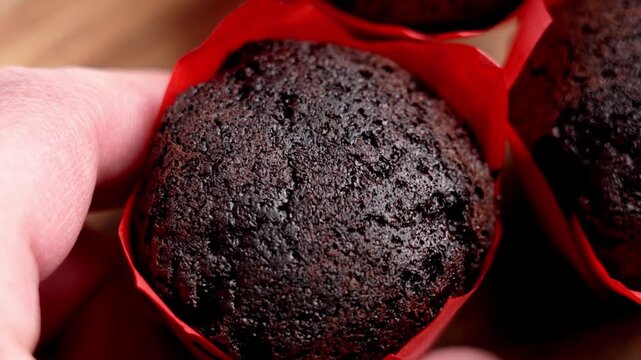 Close-up of a person hand picking up a tempting chocolate muffin in a red wrapper. Macro texture of porous cocoa sponge cake on wooden table
