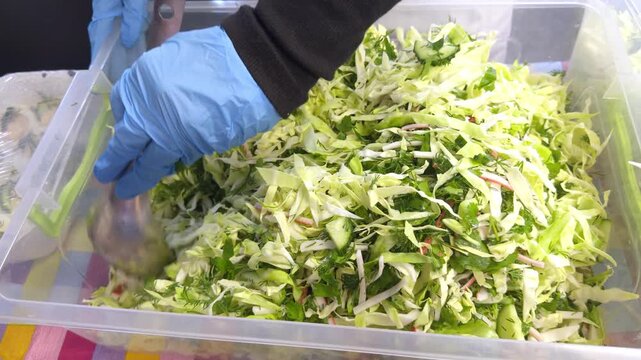 Cook in gloves mixing cabbage salad in a large plastic container in a canteen