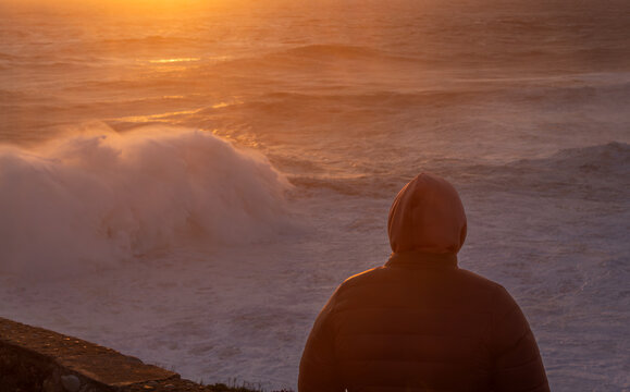 Mujer de espaldas con chaqueta y capucha contemplando grandes y turbulentas olas del mar durante el atardecer