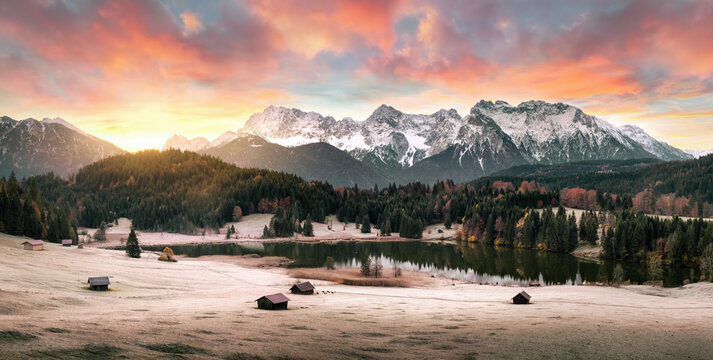 Colorful dramatic sunrise with mountains, a lake and forests in the Alps in winter, with cabins on a frozen meadow