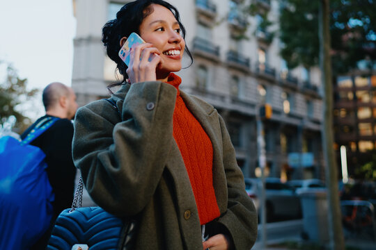 Hispanic young lady phoning on urban background, talking on mobile phone with cheerful expression, autumn street style, international roaming service, wireless communication, real people.