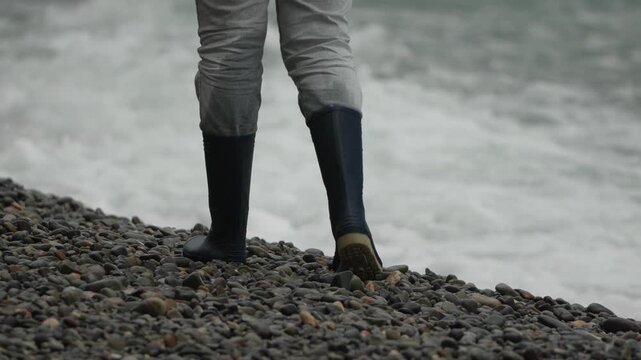 Boots, pebbles, ocean. Person wearing tall rubber boots walks down steep shingle beach picking up a stone near rough sea waves.
