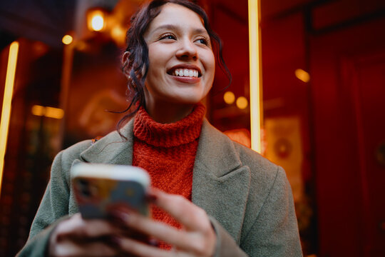 Cheerful mixed race female traveler using smartphone in city downtown, urban exploration, mobile map application, 5G connection, digital connectivity, lifestyle photography, authentic smile.