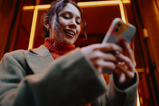 Smiling mixed race female influencer browsing content on smartphone at night, mobile application usage, positive attitude, 5G wireless internet, digital nomadic lifestyle, city leisure, happy.