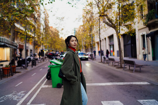 Multiracial woman in olive coat looking back while crossing street, young female traveler in autumn city setting with soft bokeh background, authentic representation of urban exploration.