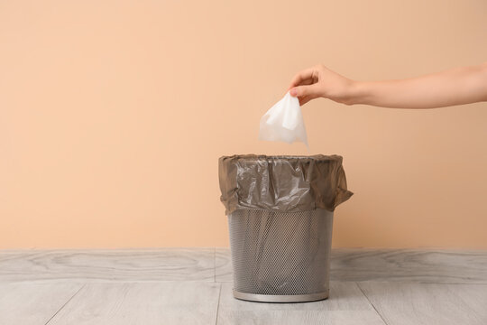 Female hand throwing used tissue into trash bin near beige wall