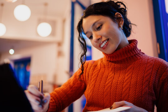 Close up of happy south asian woman using digital tablet and stylus in cafe, young female student smiling while studying online in evening, authentic representation of tech savvy professional.