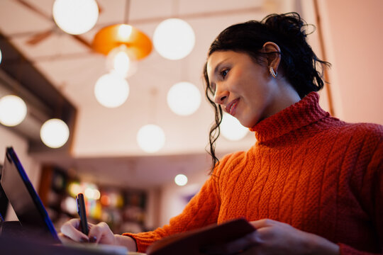 Creative young hispanic woman using electronic stylus pen on tablet screen while taking notes in journal, distance education concept, student preparing for exam in cafe, authentic representation.