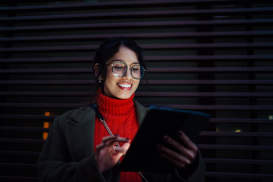 Happy multiracial female freelancer working on tablet computer in urban nighttime setting, scrolling touchscreen application, professional IT developer, creative digital nomad lifestyle.