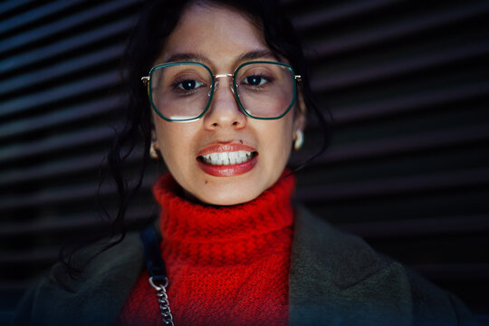 Portrait of smiling mixed race woman in green glasses and red sweater looking at camera, authentic representation of young professional, positive attitude and confidence, urban nighttime.