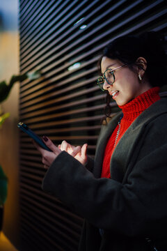 Focused multiracial woman in stylish green coat using digital tablet at night in city, creative designer scrolling mobile application, electronic communication, remote work concept.