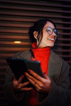 Happy mixed race woman looking away while holding tablet device outdoors at night, ambitious student dreaming about future success and career growth, digital nomad lifestyle.