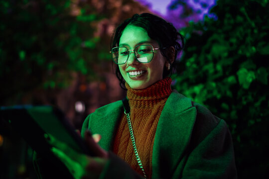 Cheerful multiracial woman using tablet computer outdoors under green light, scrolling content and smiling, professional ethnic representation, tech savvy millennial, city night life.