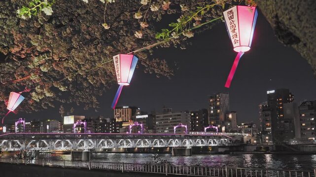 Traditional lanterns hang under cherry blossoms at night. The Sumida River and an illuminated bridge create a cinematic urban scene in Tokyo.