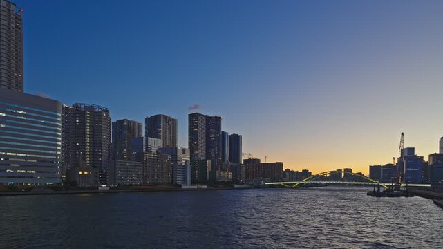 A serene evening view of the Tokyo waterfront during twilight. The calm river reflects the fading light as the city transition into night. Modern skyscrapers line the horizon, and a distinct arch brid