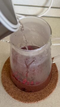 Glass pitcher with frozen currants and cranberries, boiling water poured, steam rising on tempered glass, berries releasing color