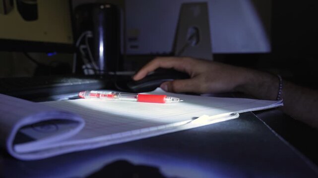 a person writing in a lined notebook with a red pen on a desk under low light, creating a focused indoor study and productivity atmosphere.