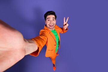 Naklejka premium Happy young man in orange suit taking a high-angle selfie with peace sign on purple background, colorful fashion portrait and playful studio mood