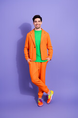 Fototapeta premium Smiling young man in bright orange suit and green shirt standing against purple studio background, colorful fashion portrait