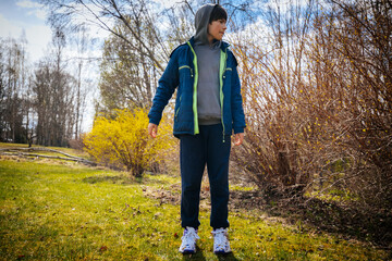 A boy in a hooded jacket stands on green grass near leafless bushes in a sunny spring park with a natural setting. © Jolanta