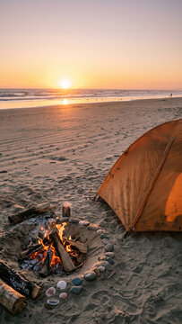 Warm campfire burning on a sandy beach next to an orange camping tent at sunset.
