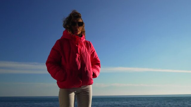 Woman sea jacket stylish young woman in pink puffer coat stands by the ocean enjoying a bright and sunny windy day