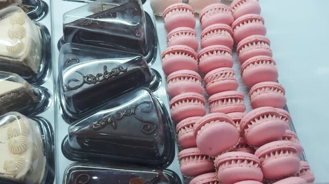 Assorted decadent pastries and confectionery in a refrigerated glass display, neatly arranged chocolate cake slices and rows of pastel pink macarons showcasing a tempting variety of sweet treats