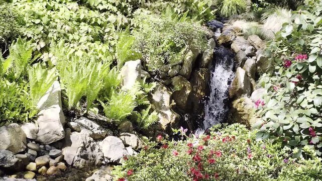 Slow motion garden waterfall over rocks with flowers, greenery and clear water in peaceful natural setting. Relaxing mobile footage in 4K Dolby Vision HDR.