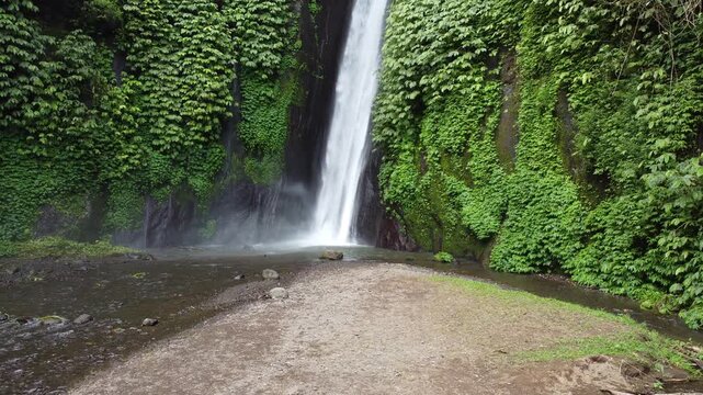 Cinematic aerial drone hovering at the base of Munduk Waterfall with tropical vegetation and flowing water, Bali, Indonesia.