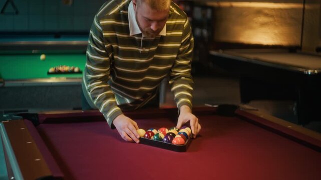 Tilt down shot of young adult man racking pool balls on red felt table using triangle while getting ready to play billiards at entertainment venue