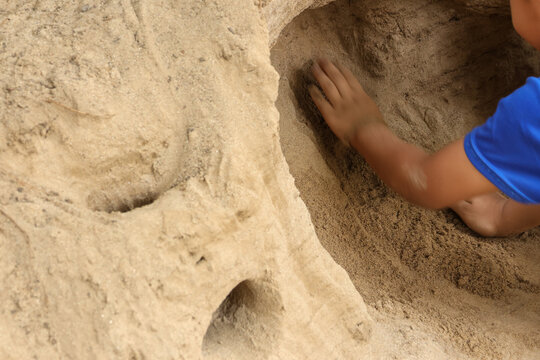 Curious playful child wearing blue shirt digging deep hole in large sand mound creating tunnel with bare hand feeling engaged in outdoor summer activity at beach
