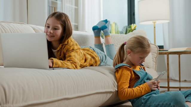 Two Caucasian kids girls modern technology addict generation looking in laptop and mobile phone smartphone sisters daughter siblings laughing enjoying using gadgets devices overuse online apps at home