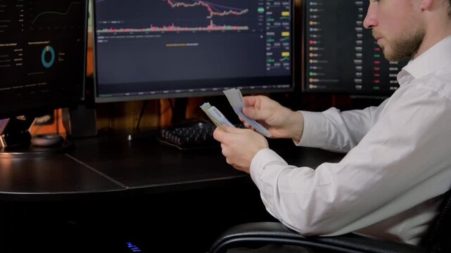Man counts cash while analyzing stock market data on multiple monitors in a dimly lit room