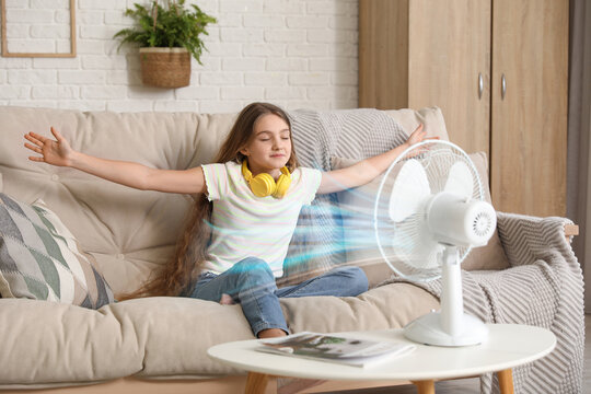 Young girl sitting on sofa with blowing electric fan in living room