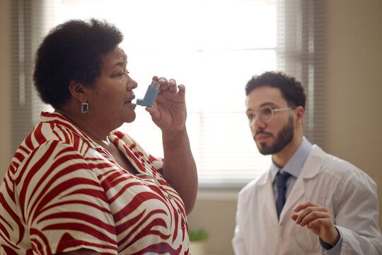 Black middle aged woman using inhaler during medical consultation while male doctor observing respiratory treatment. Ideal for asthma care, chronic disease management, patient education marketing