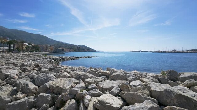 Wide angle shot of a sunlit rocky coastline meeting tranquil blue sea, with distant marina, hills and Mediterranean architecture, crisp daylight and vibrant natural colors. (198 characters)