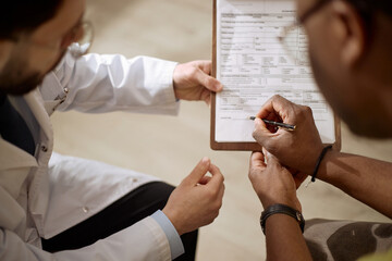 Naklejka premium Close up of Black man signing medical form while male doctor guiding paperwork during consultation. Useful for healthcare consent, patient intake, insurance, treatment planning marketing