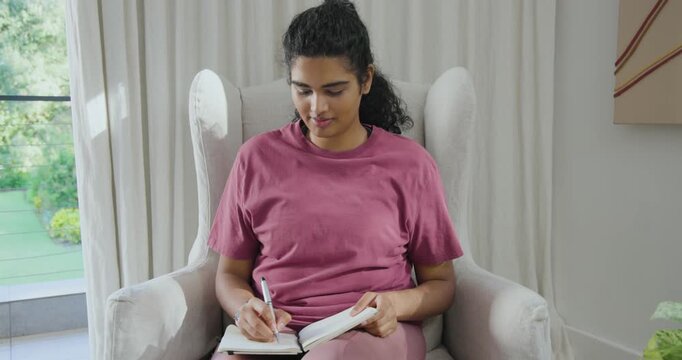 Indian woman reaching, settling back, journaling in journal with pen in magenta tee at window
