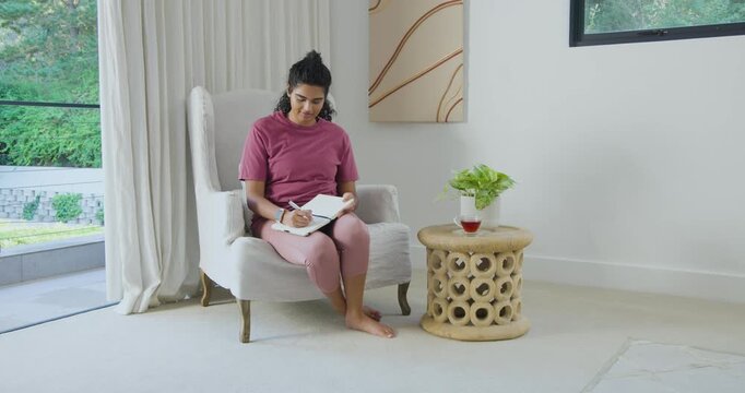 Woman sitting in armchair at home writing notebook with pen, pausing, thinking and looking outside