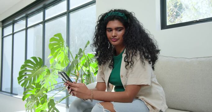 Adult woman tapping phone, checking texts, rubbing eyes on sofa by Monstera window, copy space