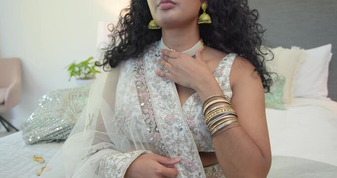 Adult Indian woman adjusting dupatta and bangles on bed, straightening pearl choker for shoot