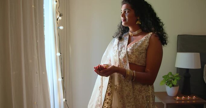 Indian woman in gold lehenga holding lit diya, lifting gaze to window, following daylight, lights