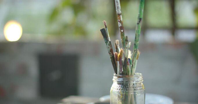 Camera is moving upward, pulling focus and revealing painted brushes in glass jar on wooden table