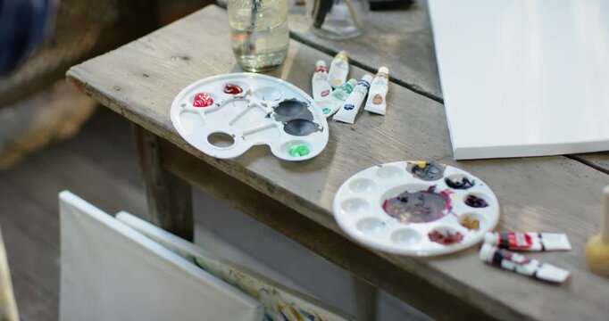 Camera is shifting right, revealing palettes, tubes, jar, canvas on bench, showing ready setup