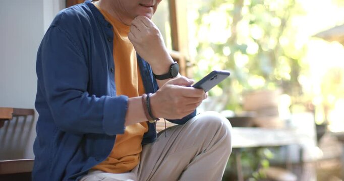 Senior Asian man orange shirt scrolling phone after alert, reading message, glancing toward garden