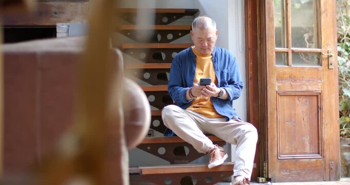 Senior Asian man sitting on wood stairs by panel door, in mustard sweater scrolling smartphone