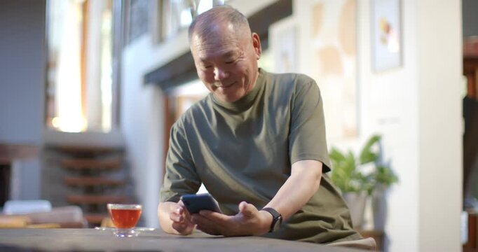 Senior Asian man scrolling smartphone, reading funny message, laughing and lifting tea at low table